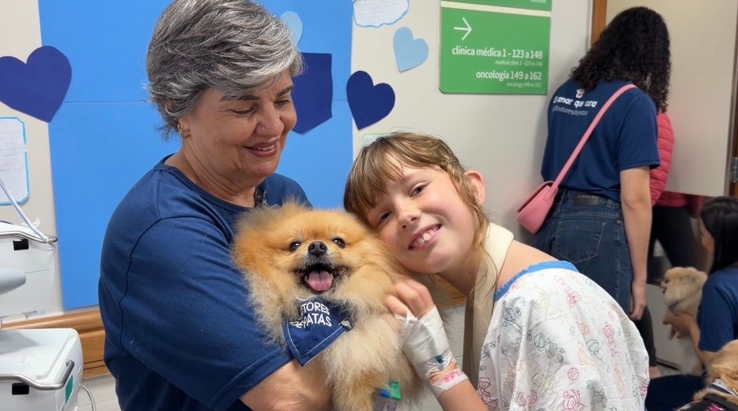 Foto mostra criança sorrindo ao interagir com cachorro, que está no colo da tutora.